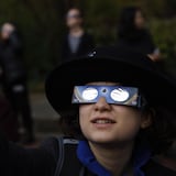 Un enfant portant des lunettes pour l'éclipse solaire regarde vers le ciel et pointe son doigt dans cette direction.