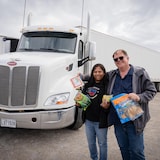 Un homme et une femme avec des produits alimentaires dans les bras devant un camion et une remorque blanche.