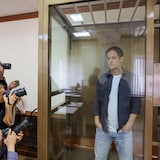 Evan Gershkovich est debout dans une prison de verre alors que des photographes le prennent en photo.