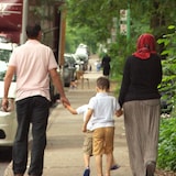 Un homme marchant à gauche sur le trottoir et portant un t-shirt beige tient la main d'un enfant, qui marche au centre du trottoir. Une femme portant un hijab rouge, marche à droite sur le trottoir et tient la main d'un second enfant.
