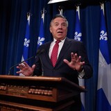 Isang lalaki nasa podium habang may mga watawat ng Quebec sa kanyang likuran.