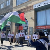 أشخاص يحملون لافتات وامرأة تحمل العلم الفلسطيني أمام مبنى.