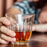 A person resting their head on their arm on a table while still grabbing a glass of liquor.