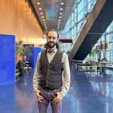 Español : Retrato de un hombre con barba portando un chaleco, manos en los bolsillos, en un vestíbulo luminoso.
Français : Portrait d'un homme barbu portant un gilet, debout, mains dans les poches, dans un hall lumineux.