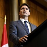رئيس الحكومة الكندية جوستان ترودو متحدثاً في مؤتمر صحفي، ونراه واقفاً خلف منبر وخلفه جزء من علم كندا.