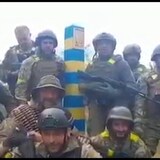 Plusieurs hommes armés en treillis militaires sont debout ou agenouillés près d'un poteau peint en bleu et jaune. 