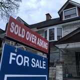 Une affiche de vente immobilière, sur laquelle on peut lire « Sold over asking » (vendu au-dessus du prix demandé), devant des maisons en rangée.