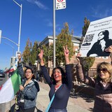 Tres mujeres en la acera muestran signos de paz con sus manos. Una sostiene un póster de Mahsa Amini y otra una bandera iraní.