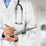 Un médecin dans un couloir d'hôpital s'apprête à écrire sur un document. Il porte un stéthoscope autour du cou.