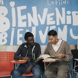 Deux hommes lisent sous une fresque murale "BIENVENIDOS BIENVENUE" dans un centre d'accueil.