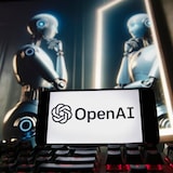 Un robot humanoide futurista mirando su reflejo en un espejo en una pantalla. Delante de la pantalla, un teclado y un celular con el logotipo de OpenAI.