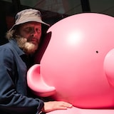 Philippe Katerine regarde la caméra. Il est accoté sur la tête ronde de sa grosse sculpture rose.