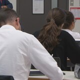 Des étudiants sont assis à des tables, ordinateurs devant eux. Ils regardent vers l'avant ou prennent des notes. 