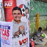 na mujer indígena vende comida junto a un cartel electoral del candidato presidencial Rodrigo Paz Pereira del Partido Demócrata Cristiano en La Paz, Bolivia, el 17 de octubre de 2025. Elle est assise sur un trottoir.