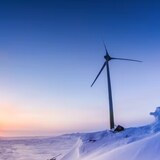 The sun goes down on a snowy landscape behind a wind turbine. 