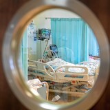 Une personne couchée dans un lit d'hôpital, photographiée à travers la fenêtre circulaire d'une porte fermée.