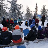 Groupe d'enfants habillés avec vêtements d'hiver assis autour d'un feu dehors avec tente et arbres recouverts de neige, le 30 novembre 2022, à Yellowknife.
