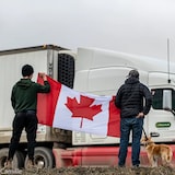 Dalawang lalaki ang may hawak na watawat ng Canada habang dumadaan ang mga trucker.