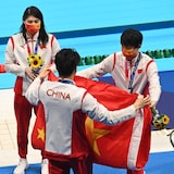 四名身着运动服的运动员站在游泳池边，展开一面中国国旗。