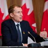 Le ministre parle assis à une table lors d'une conférence de presse avec des drapeaux du Canada en toile de fond.