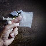 Une main qui tient de l'argent et de la poudre d'héroïne dans un sac. Une seringue, un médicament, une cigarette et un briquet sont déposés sur une table à l'arrière-plan.