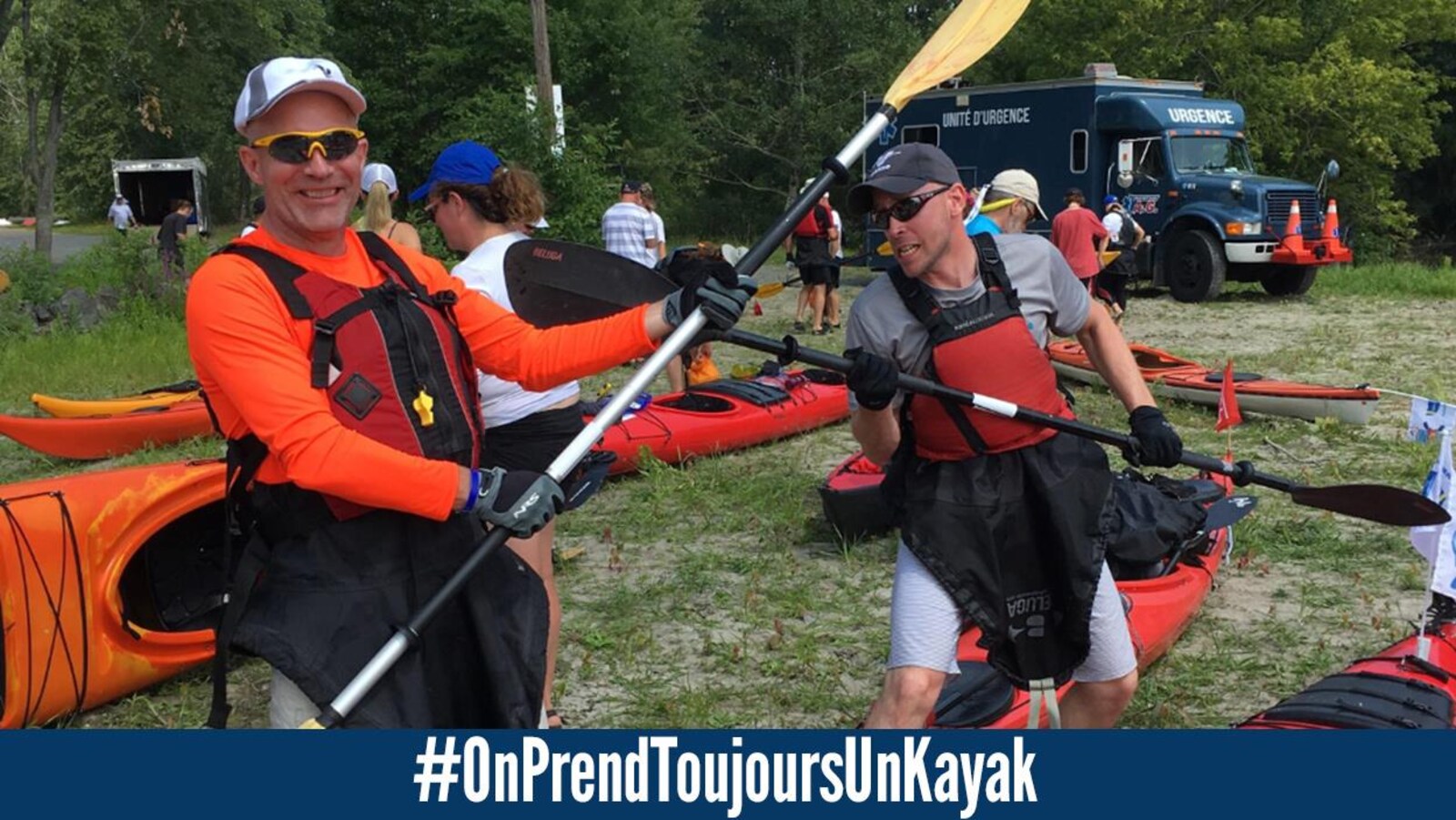 Deux hommes portant des vestes de sauvetage, des casquettes, des lunettes fumées et des jupettes de kayak, font semblant de se battre avec les pagaies en guise d'épée. Derrière, on voit d'autres kayakistes, des kayaks sur le sol et un véhicule d'urgence.