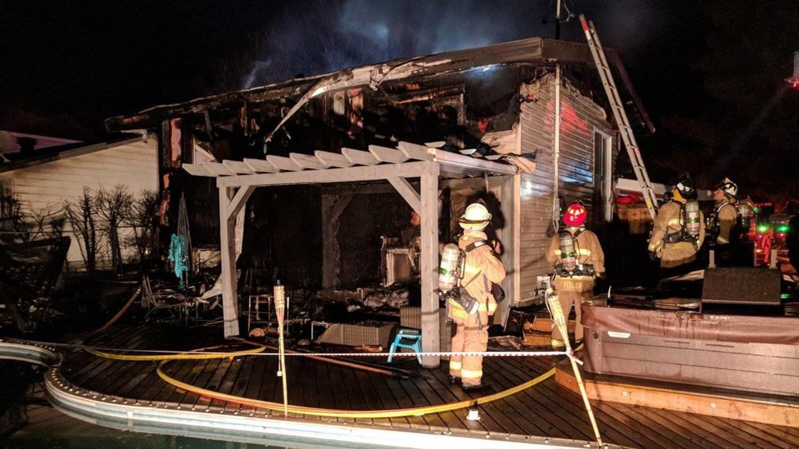 Des pompiers sur une pergola tentent d'éteindre un feu dans une maison. Une piscine et un sauna au premier plan.