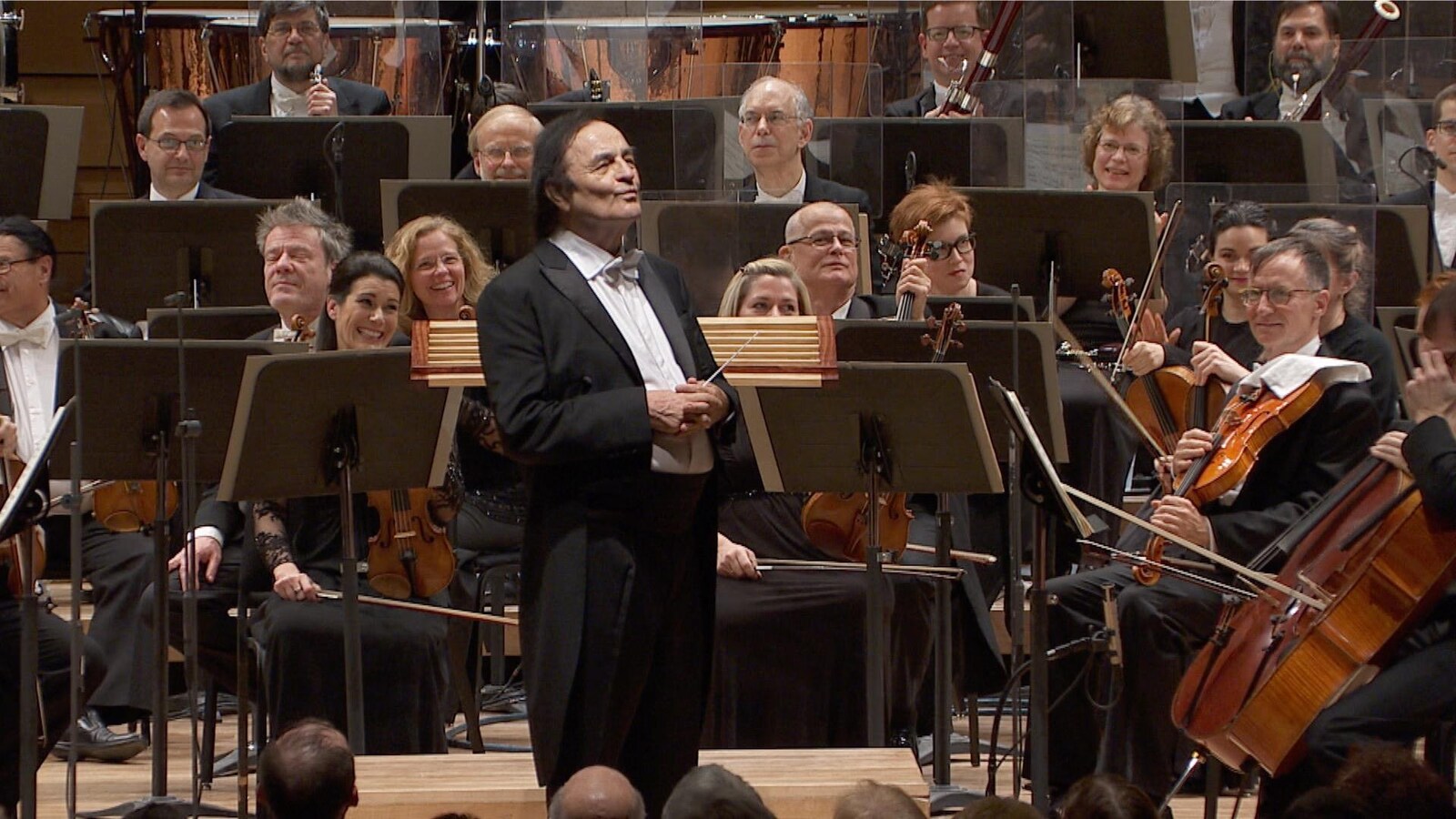 Charles Dutoit est debout sur scène, au milieu d'un orchestre.