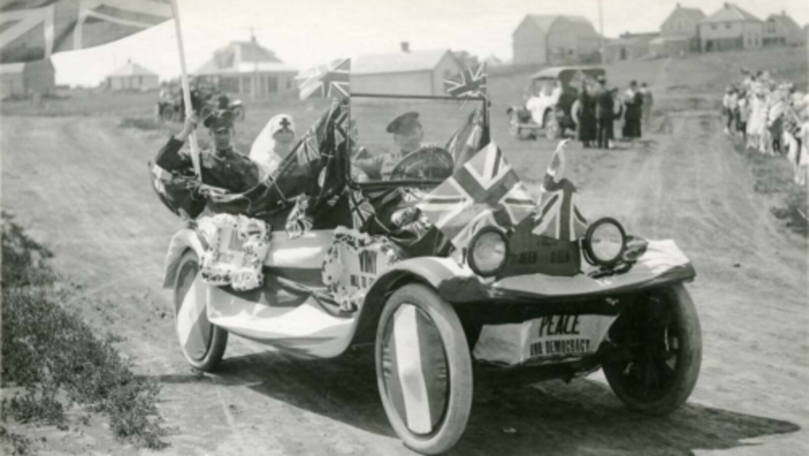Une vieille voiture d'antan sans toit (1919) avec des soldats et une infirmière à l'intérieur et des drapeaux partout sur la voiture. C'est écrit ''paix'' devant la voiture.