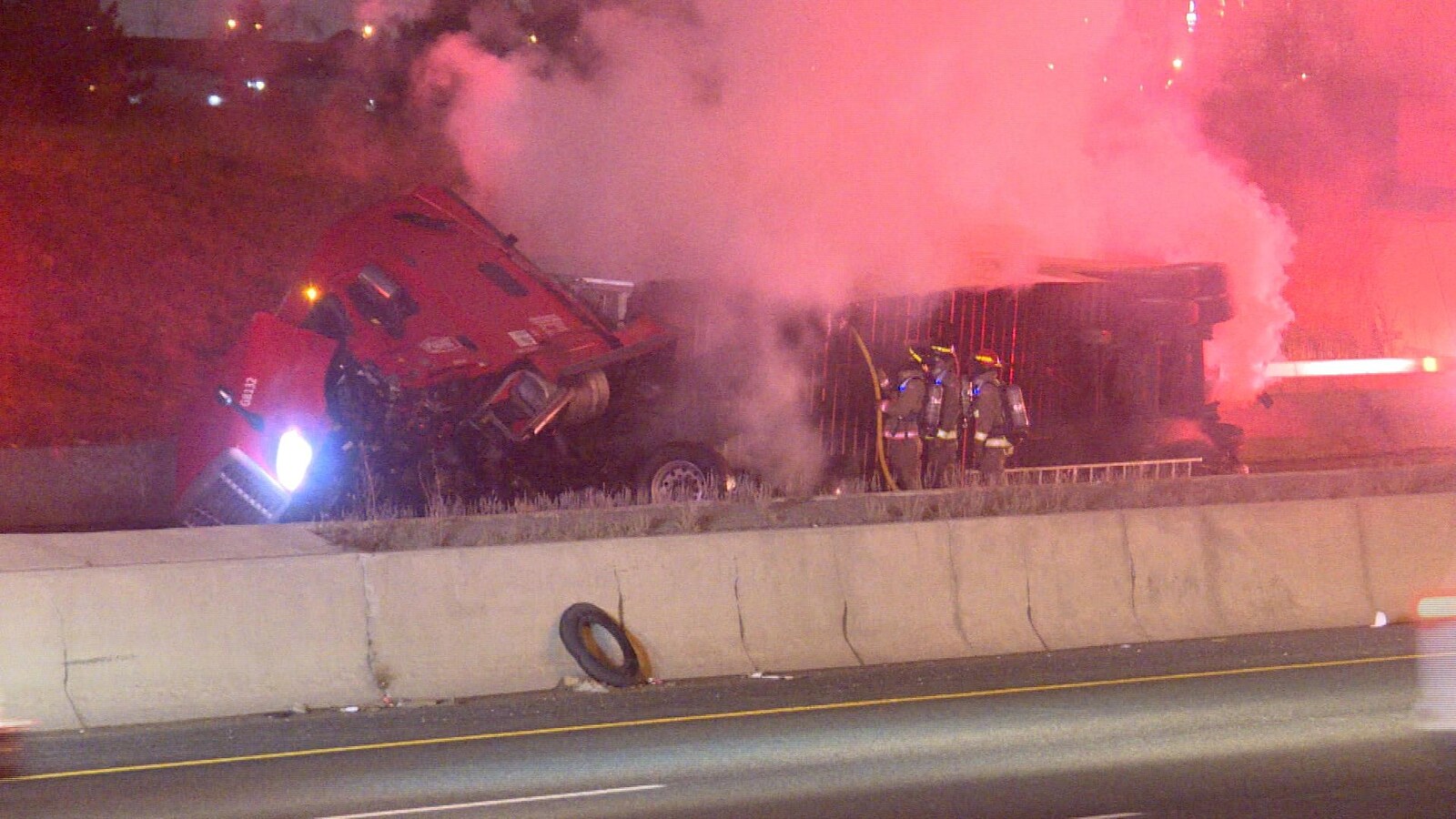 Photo de fumée qui se dégage d'un camion en feu; des pompiers arrosent sous la remorque renversée du camion.