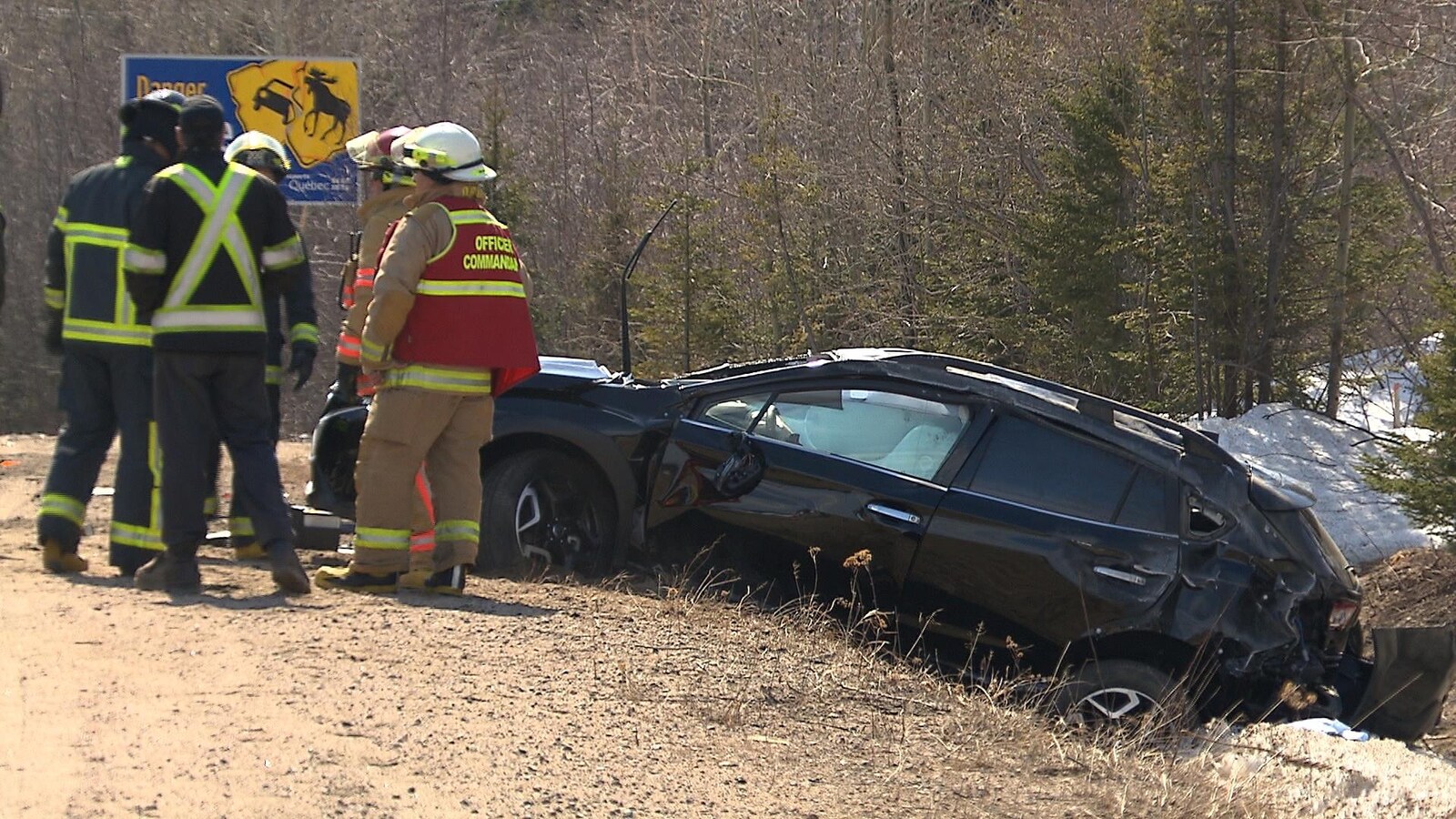Une auto noire, accidentée, est dans le fossé et des pompiers se tiennent tout près.