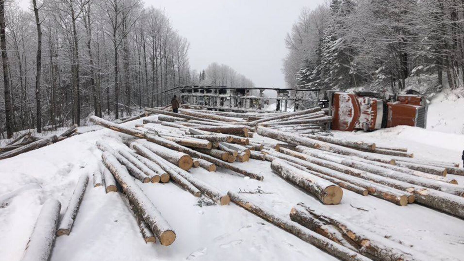 Le chargement de bois d'un camion-remorque jonché en plein milieu de la chaussée avec le camion renversé sur le côté perpendiculaire à la route.