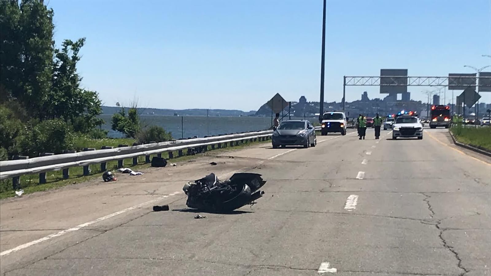 Scène de l’accident. On aperçoit la moto accidentée, couchée sur le côté. À l’arrière-plan, on distingue des véhicules d’urgence stationnés sur la voie est de l’autoroute Dufferin-Montmorency, laquelle est bordée par le fleuve Saint-Laurent. Au loin se profile le centre-ville de Québec.