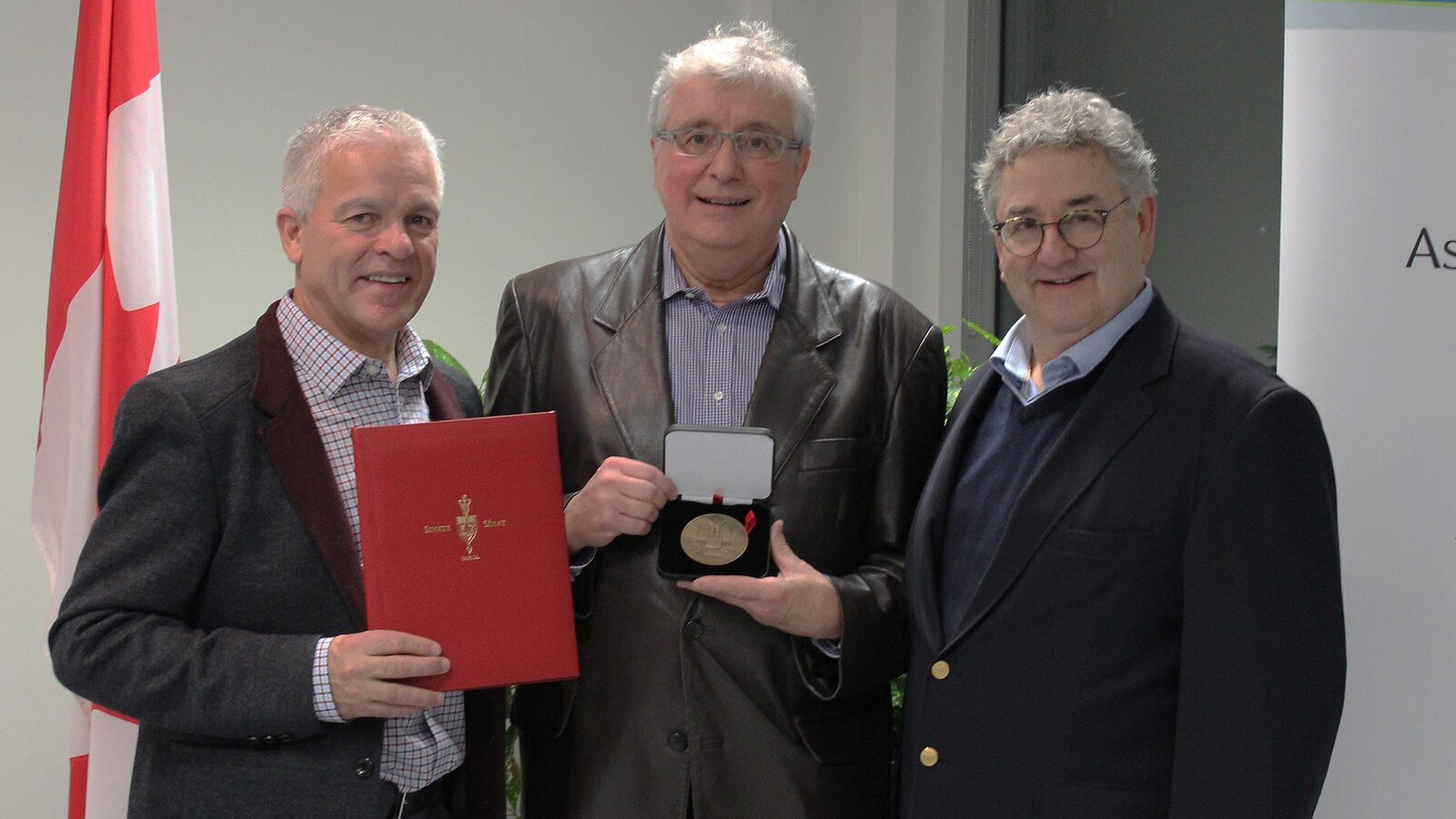 Trois hommes prennent la pose aux côtés d'un drapeau du Canada. L'un d'eux tient une médaille du Sénat qu'il s'est vu décerner.