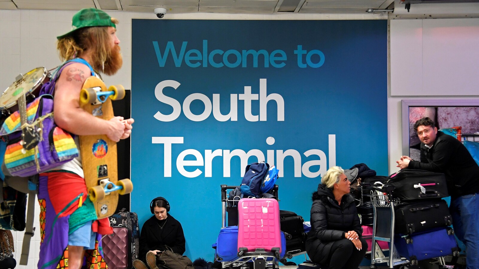Un homme barbu avec une planche à roulettes, un banjo et des vêtements très colorés passe devant des passagers entourés de valises.