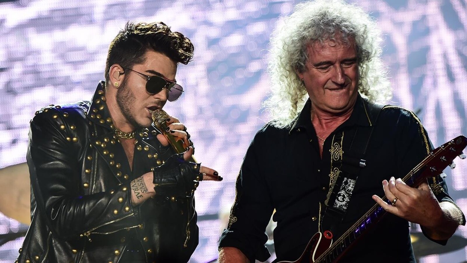 Le chanteur Adam Lambert et Brian May, du groupe Queen, sont sur scène et interprète un des succès du groupe devant un grand écran illuminé.