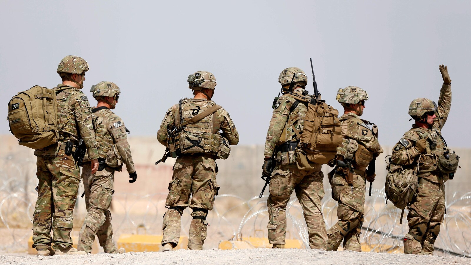 Six soldats américains marchent sur un sol terreux, le long d'une clôture de barbelés.