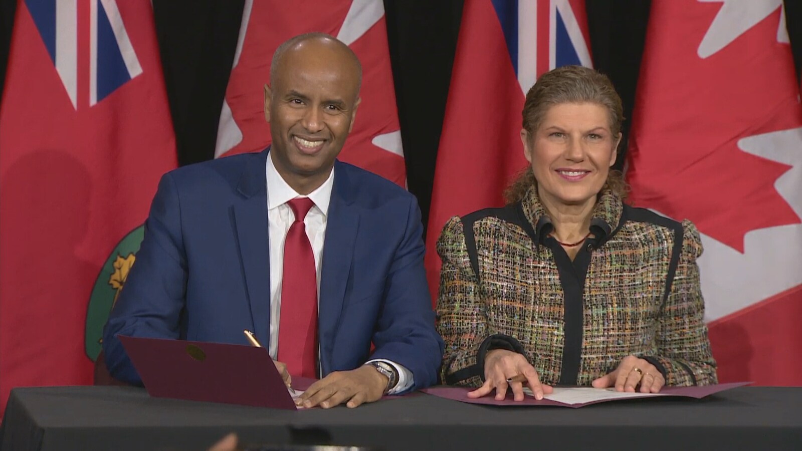 Les deux ministres sont assis à une table, crayons à la main, prêts à signer l'entente. Ils regardent la caméra en souriant.
