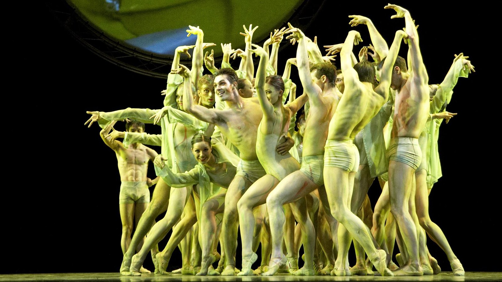 Une troupe de danseurs, habillés et maquillés dans les tons de jaune et de vert, sont rassemblés sur scène.