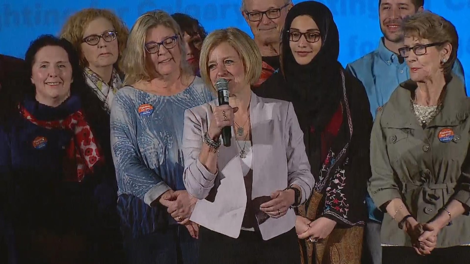 Une femme parle dans un micro. Il y a du monde derrière elle qui l'écoute. Ils sont tous debout.