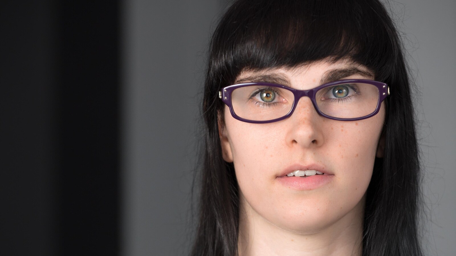 Portrait en couleur de l'auteure Alex Thibodeau sur fond gris : elle porte des lunettes mauves, un chandail gris, des cheveux bruns lâchés et une frange.