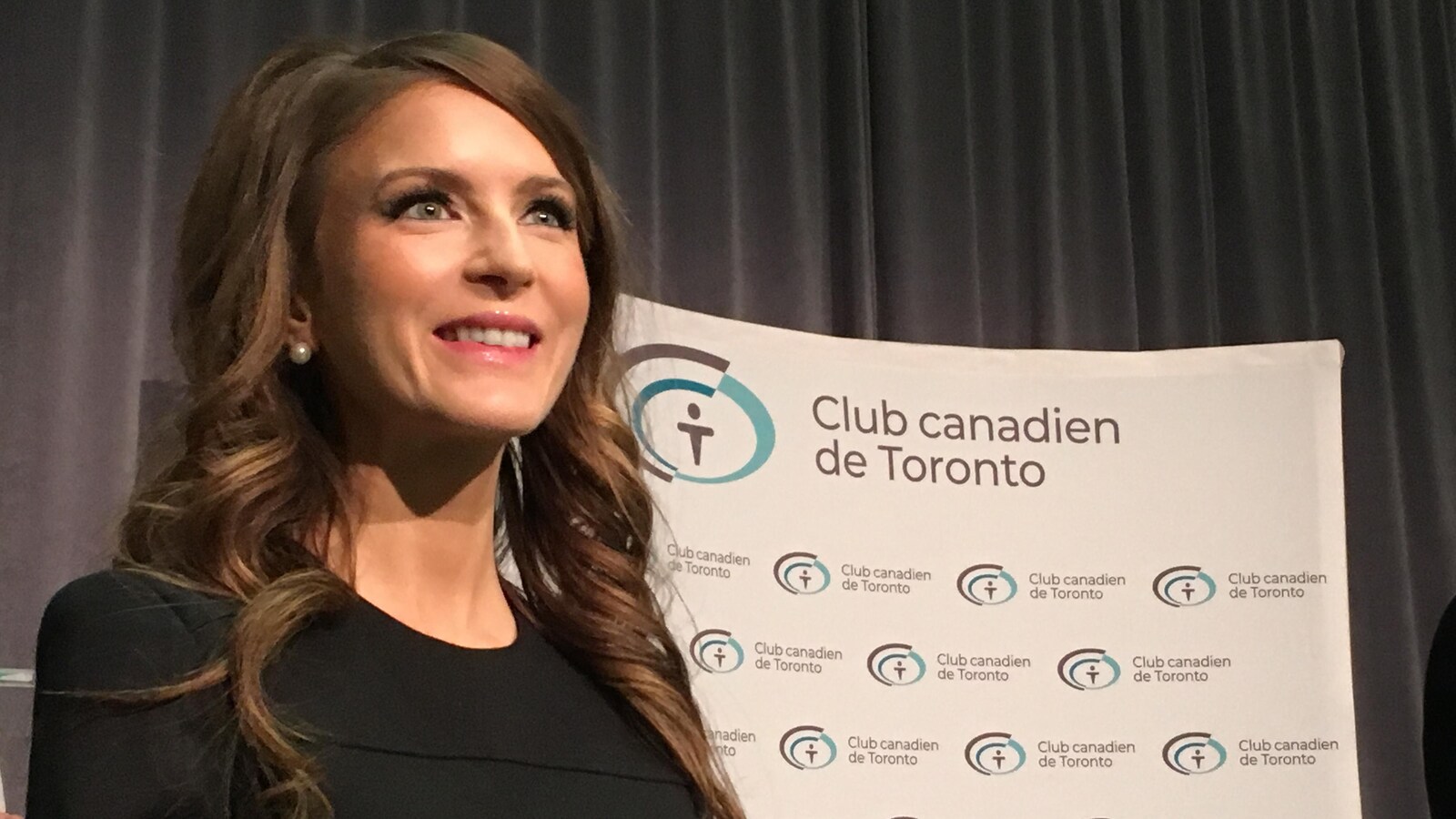 Une femme aux cheveux longs vêtue d'une robe noire sourit devant une affiche qui dit « Club canadien de Toronto ».