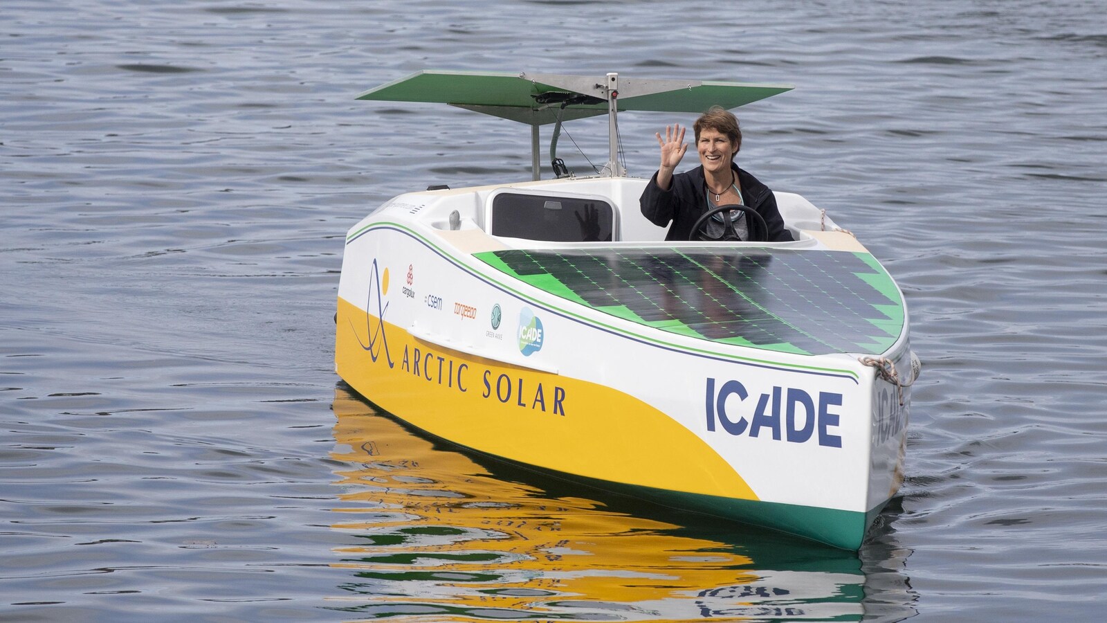 Une femme fait un signe de la main alors qu'elle est à la barre d'un petit bateau, nommé Icade, sans voile, ne fonctionnant qu'à l'énergie solaire. Des panneaux solaires recouvrent tout l'avant du bateau.