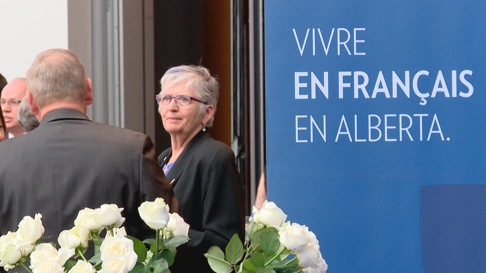 Une bannière où se lit «Vivre en français en Alberta», à droite, avec la députée Annie McKitrick qui écoute un interlocuteur, vu de dos, avec des roses blanches au bas de l'image.