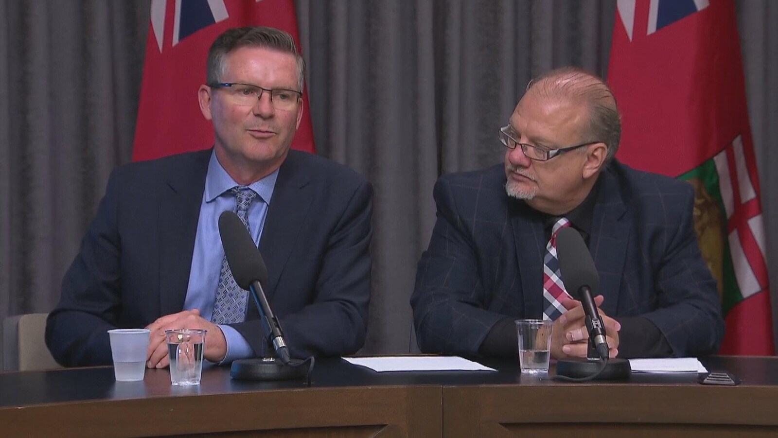 Deux hommes en complet sont assis à une table devant des drapeaux manitobains et ont des microphones devant eux.