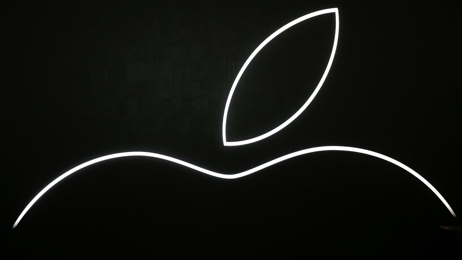 Une photo montrant le haut du logo d'Apple en blanc sur fond noir. Dans le bas de l'écran, les têtes de spectateurs devant l'écran sont visibles dans la pénombre.