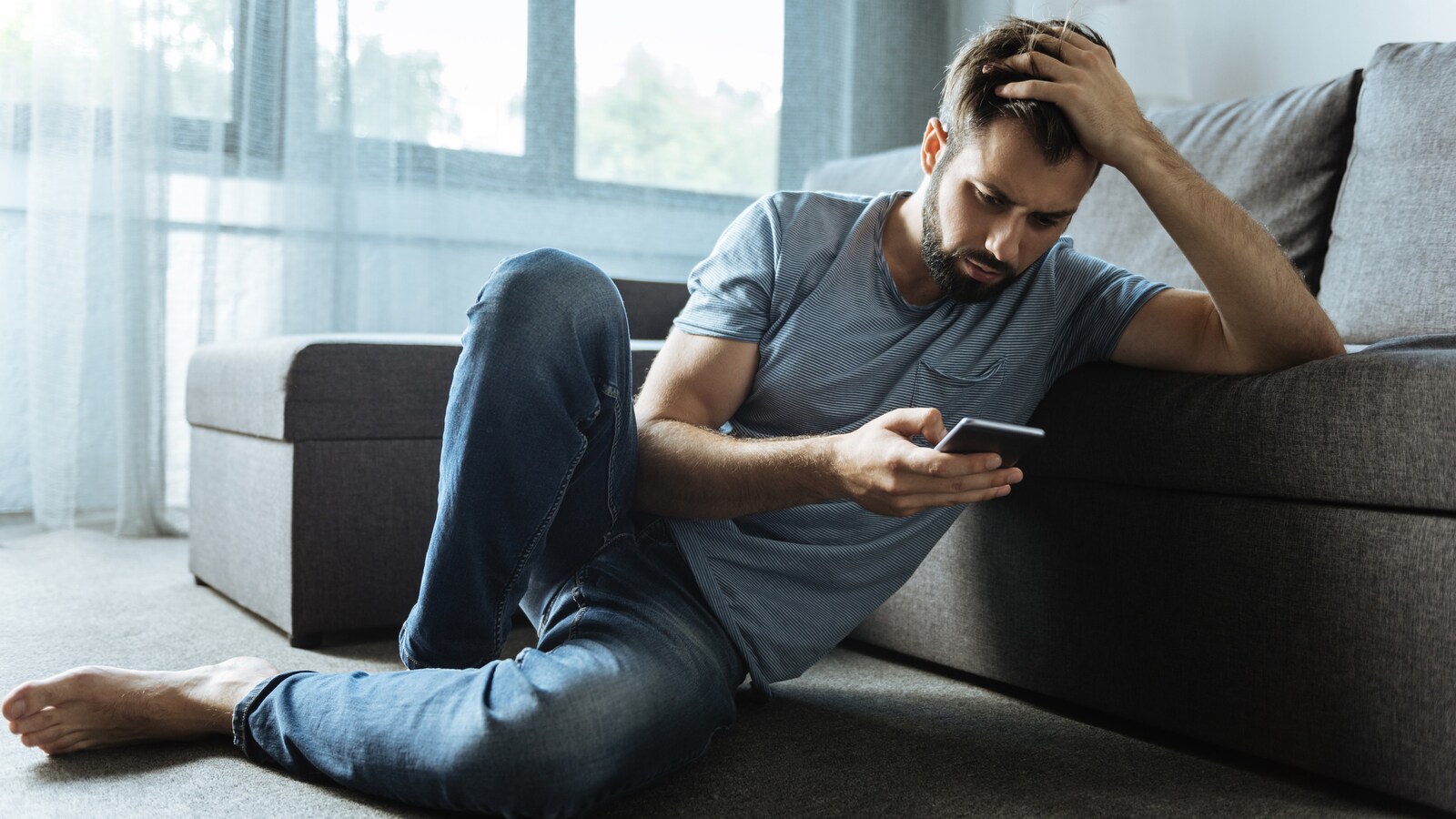 Un homme triste consulte son téléphone. Il est assis sur le tapis de son salon consulte son cellulaire.