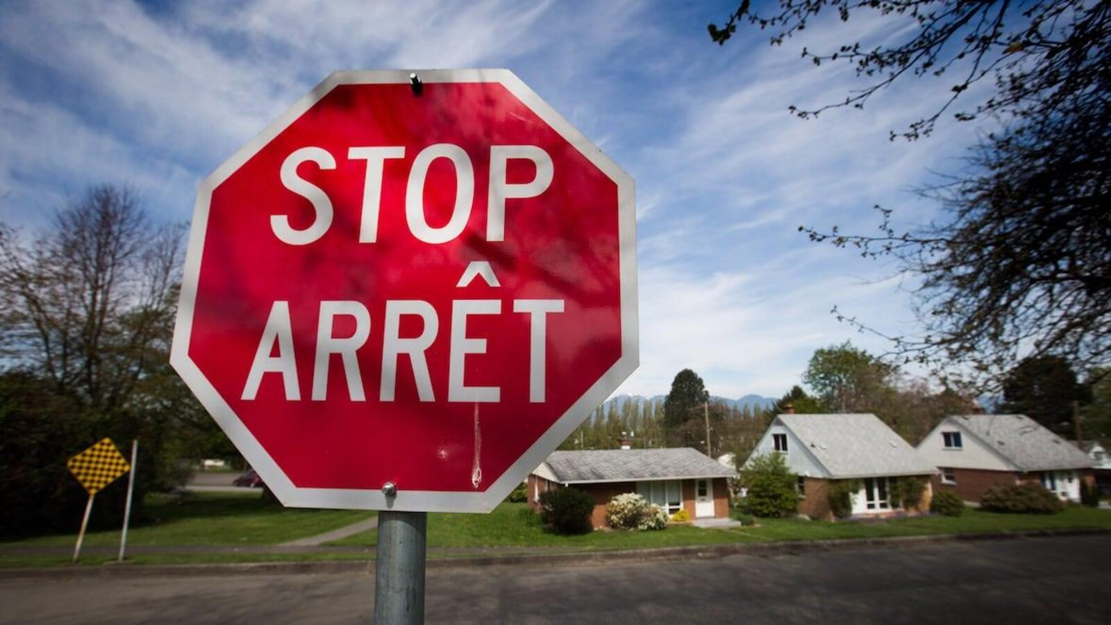 Un panneau arrêt-stop bilingue rouge et blanc de forme octogonale se trouve au premier plan. Il y a des maisons au second plan.