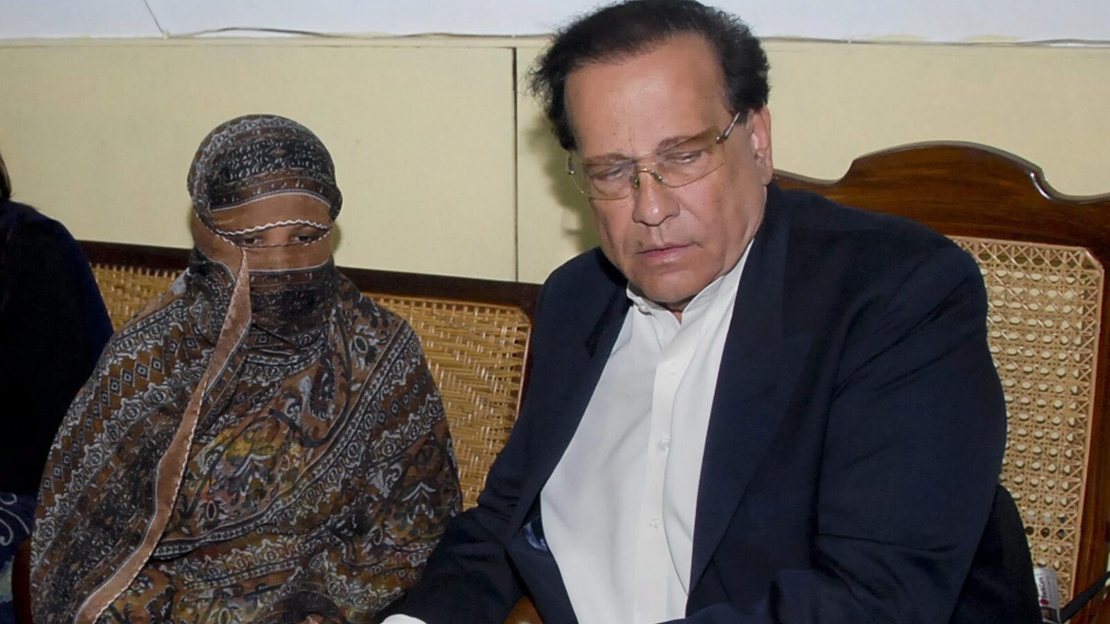 Une femme recouverte d'un voile à motifs couvrant tout sauf ses yeux est assise aux côtés d'un homme portant lunettes et veston.