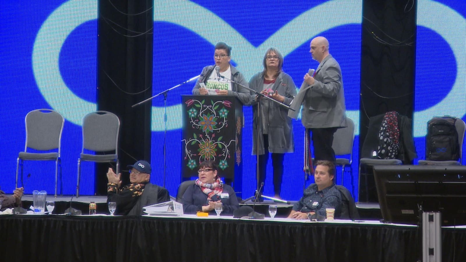 Trois personnes sur un podium avec le drapeau métis en arrière-plan.